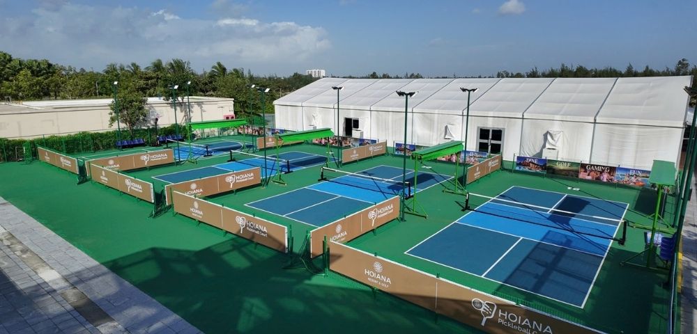 Tennis complex with multiple courts enclosed by brown barriers under a clear blue sky. A large white tent and palm trees are visible in the background.