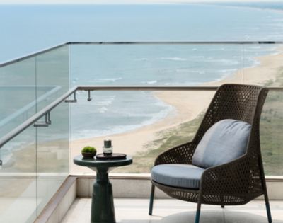 Cozy balcony with a wicker chair and small table holding a plant and cup. Overlooks a serene beach and ocean, evoking calm and relaxation.