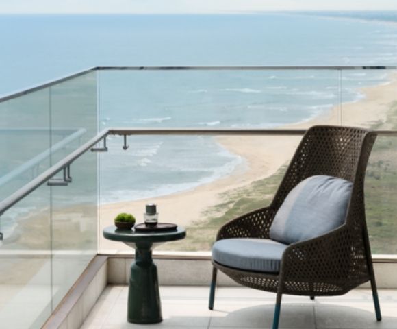 Cozy balcony with a wicker chair and small table holding a plant and cup. Overlooks a serene beach and ocean, evoking calm and relaxation.