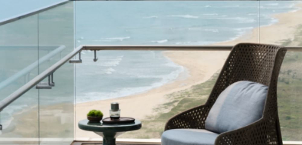 Cozy balcony with a wicker chair and small table holding a plant and cup. Overlooks a serene beach and ocean, evoking calm and relaxation.