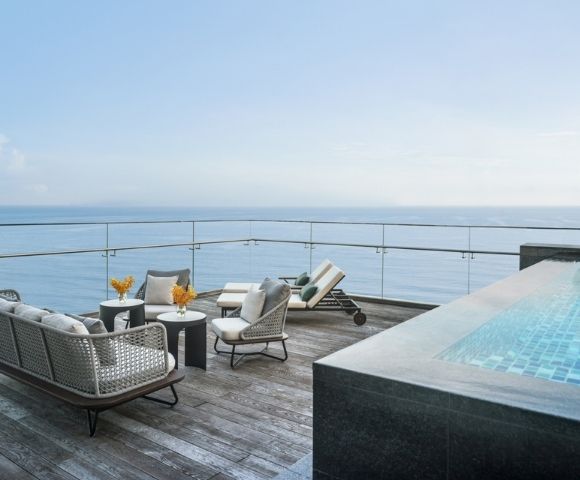 Rooftop patio with wicker furniture and a lounge chair, overlooking the ocean. Nearby, a modern infinity pool adds to the serene, luxurious ambiance.