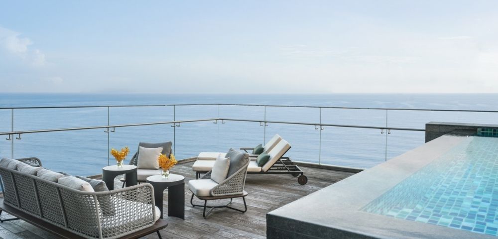 Rooftop patio with wicker furniture and a lounge chair, overlooking the ocean. Nearby, a modern infinity pool adds to the serene, luxurious ambiance.