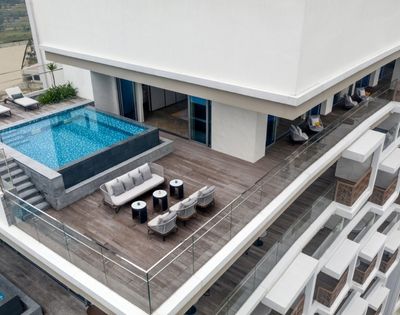 Rooftop view featuring a modern rectangular pool, wooden deck, and plush seating with tables. Glass railings offer a scenic, luxurious ambiance.