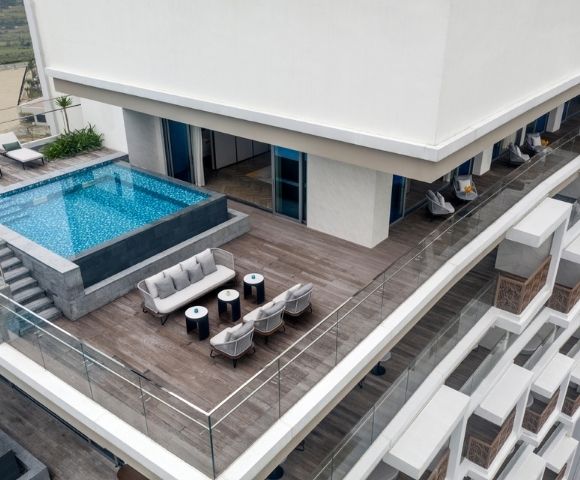 Rooftop view featuring a modern rectangular pool, wooden deck, and plush seating with tables. Glass railings offer a scenic, luxurious ambiance.
