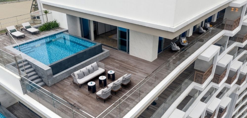 Rooftop view featuring a modern rectangular pool, wooden deck, and plush seating with tables. Glass railings offer a scenic, luxurious ambiance.