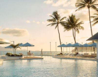 Sunlit poolside scene featuring loungers and blue umbrellas, surrounded by palm trees with an ocean view. The atmosphere is tranquil and relaxing.