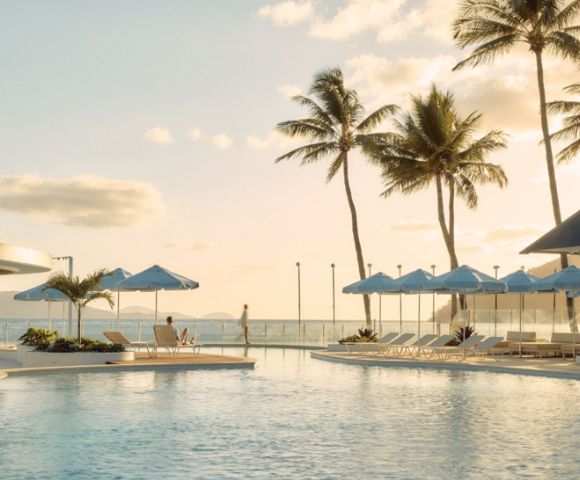 Sunlit poolside scene featuring loungers and blue umbrellas, surrounded by palm trees with an ocean view. The atmosphere is tranquil and relaxing.
