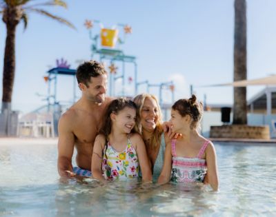 A happy family of four, two adults and two children, enjoy a sunny day in a shallow pool. Palm trees and colorful water park structures are in the background.