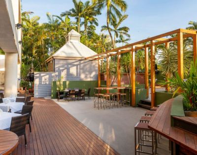 Outdoor seating area with wooden decking and stools, surrounded by lush palm trees. The atmosphere is serene and inviting under a clear blue sky.