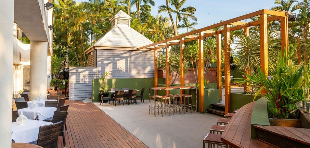 Outdoor seating area with wooden decking and stools, surrounded by lush palm trees. The atmosphere is serene and inviting under a clear blue sky.
