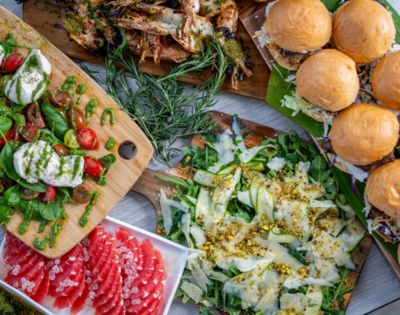 A vibrant spread of food, including burgers, fresh salads with mozzarella and basil, grilled shrimp, and sliced grapefruit, creates a feast-like atmosphere.