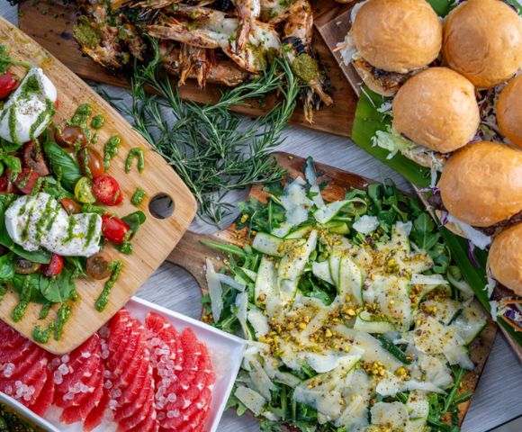 A vibrant spread of food, including burgers, fresh salads with mozzarella and basil, grilled shrimp, and sliced grapefruit, creates a feast-like atmosphere.