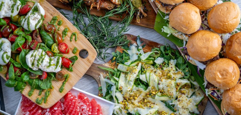 A vibrant spread of food, including burgers, fresh salads with mozzarella and basil, grilled shrimp, and sliced grapefruit, creates a feast-like atmosphere.
