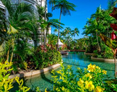 Lush tropical garden with a meandering pool surrounded by vibrant green palm trees and yellow flowers under a clear blue sky, exuding tranquility.