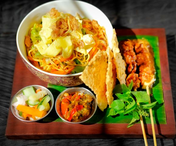 A vibrant Indonesian meal featuring a bowl of noodles with vegetables, crispy crackers, chicken skewers, and fresh herbs, accompanied by pickles and sambal in small dishes.