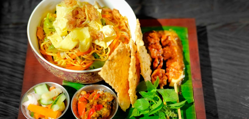 A vibrant Indonesian meal featuring a bowl of noodles with vegetables, crispy crackers, chicken skewers, and fresh herbs, accompanied by pickles and sambal in small dishes.