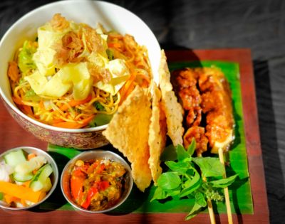 A vibrant Indonesian meal featuring a bowl of noodles with vegetables, crispy crackers, chicken skewers, and fresh herbs, accompanied by pickles and sambal in small dishes.