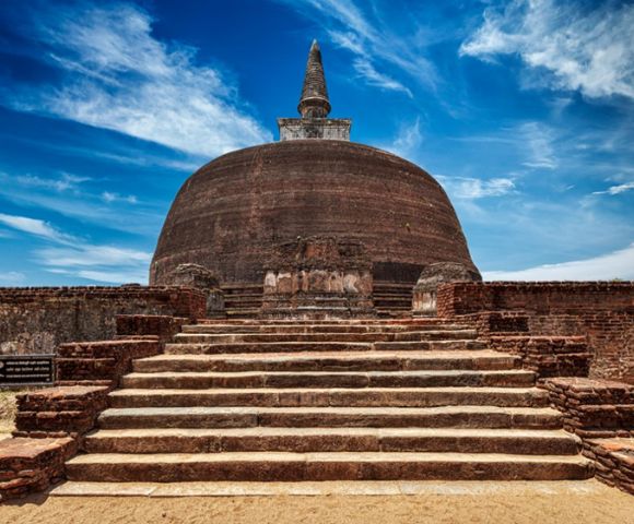 Ancient stone stupa under a vibrant blue sky with wispy clouds, surrounded by weathered brick walls. The scene conveys historical significance and tranquility.