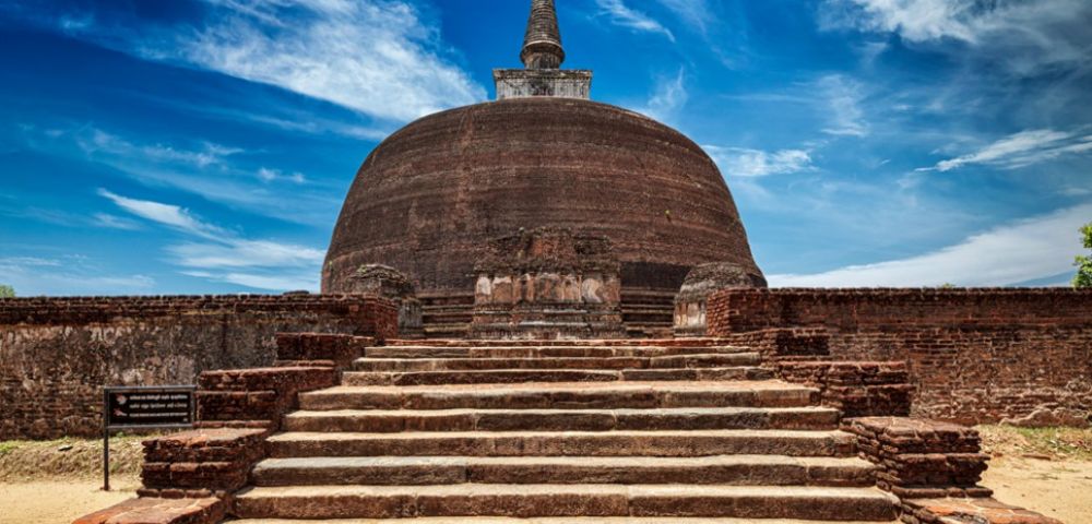 Ancient stone stupa under a vibrant blue sky with wispy clouds, surrounded by weathered brick walls. The scene conveys historical significance and tranquility.