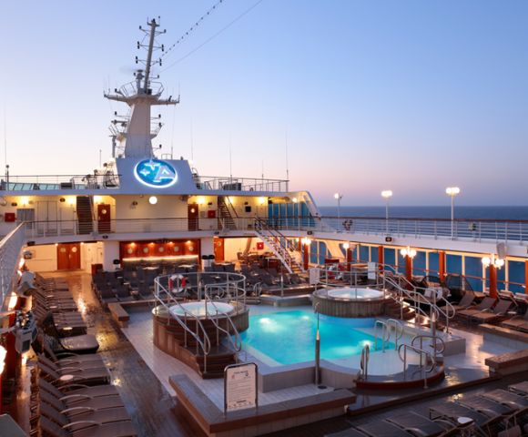 Cruise ship deck at sunset with glowing pool and whirlpools, surrounded by lounge chairs. Tranquil ocean horizon in the background.