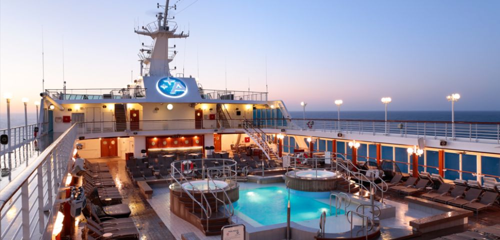 Cruise ship deck at sunset with glowing pool and whirlpools, surrounded by lounge chairs. Tranquil ocean horizon in the background.