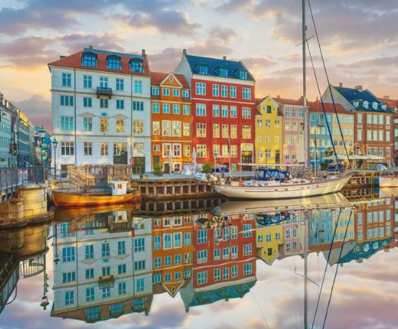 Colorful buildings are reflected in a calm canal at sunset, with a sailboat docked in front. The sky is painted with soft pastel hues.