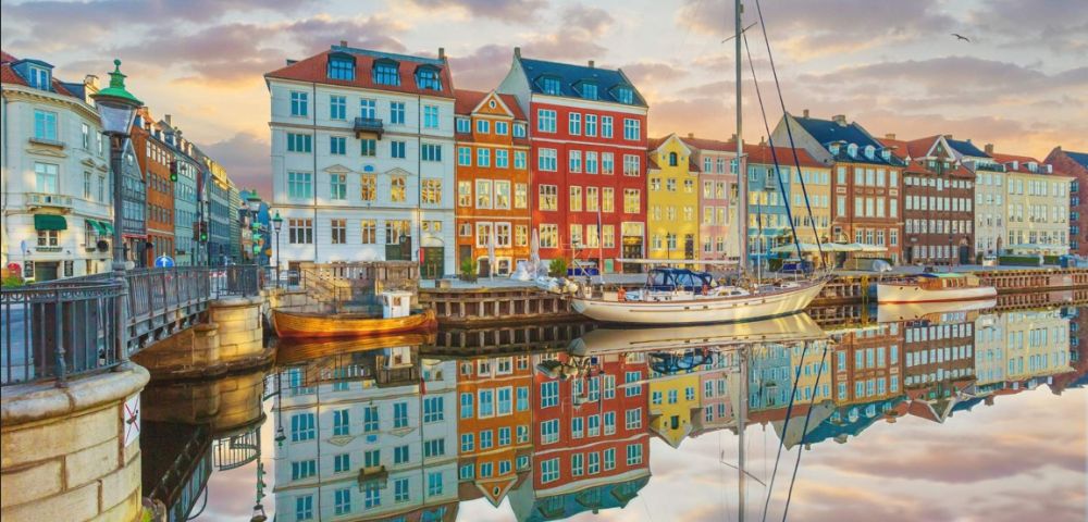 Colorful buildings are reflected in a calm canal at sunset, with a sailboat docked in front. The sky is painted with soft pastel hues.