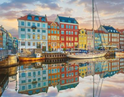 Colorful buildings are reflected in a calm canal at sunset, with a sailboat docked in front. The sky is painted with soft pastel hues.
