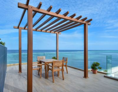 A wooden pergola on a seaside deck with a dining table and chairs. The scene conveys tranquility, with blue skies and ocean views in the background.