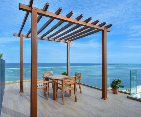 A wooden pergola on a seaside deck with a dining table and chairs. The scene conveys tranquility, with blue skies and ocean views in the background.