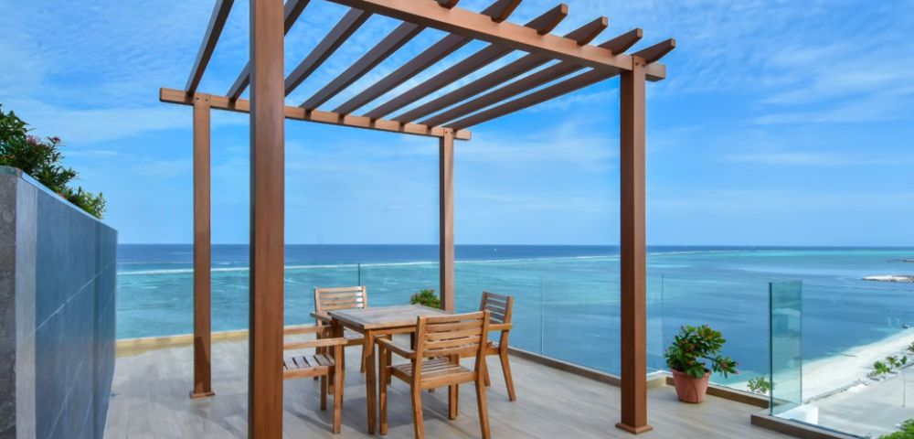 A wooden pergola on a seaside deck with a dining table and chairs. The scene conveys tranquility, with blue skies and ocean views in the background.