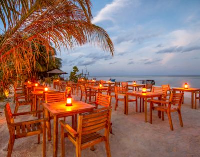 Beachside restaurant at sunset with wooden tables and chairs on sand, lit by candles. Palm tree on the left, ocean view under a cloudy sky. Peaceful atmosphere.