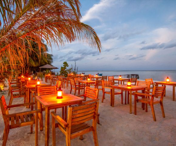 Beachside restaurant at sunset with wooden tables and chairs on sand, lit by candles. Palm tree on the left, ocean view under a cloudy sky. Peaceful atmosphere.
