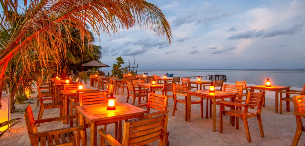 Beachside restaurant at sunset with wooden tables and chairs on sand, lit by candles. Palm tree on the left, ocean view under a cloudy sky. Peaceful atmosphere.