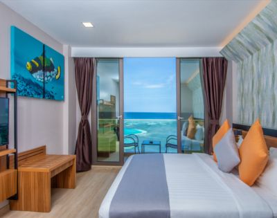 Modern hotel room with an ocean view, featuring a bed with colorful pillows, a wooden bench, and a vibrant fish artwork. Bright and inviting atmosphere.