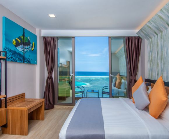 Modern hotel room with an ocean view, featuring a bed with colorful pillows, a wooden bench, and a vibrant fish artwork. Bright and inviting atmosphere.