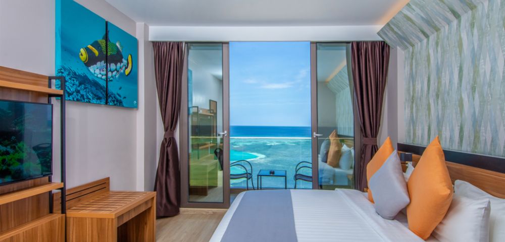 Modern hotel room with an ocean view, featuring a bed with colorful pillows, a wooden bench, and a vibrant fish artwork. Bright and inviting atmosphere.