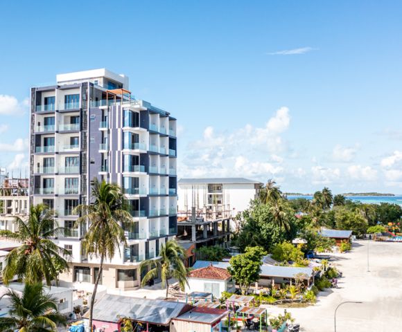 Modern multi-story building with palm trees in a tropical setting. Clear blue sky and distant ocean suggest a coastal, relaxing atmosphere.