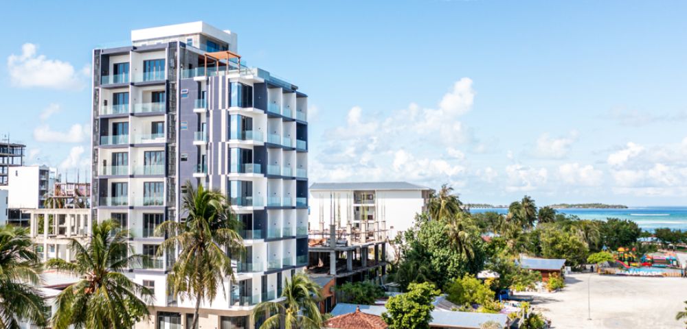 Modern multi-story building with palm trees in a tropical setting. Clear blue sky and distant ocean suggest a coastal, relaxing atmosphere.