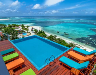 A rooftop infinity pool overlooks a vibrant turquoise ocean and sandy beach. Bright orange and green lounge chairs add a lively touch to the tranquil scene.