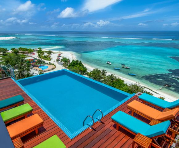 A rooftop infinity pool overlooks a vibrant turquoise ocean and sandy beach. Bright orange and green lounge chairs add a lively touch to the tranquil scene.