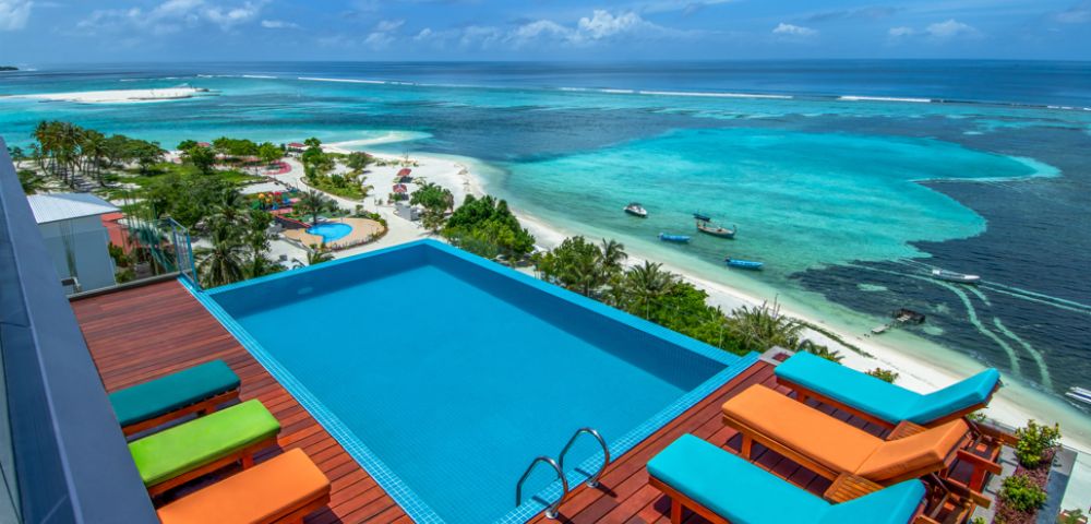 A rooftop infinity pool overlooks a vibrant turquoise ocean and sandy beach. Bright orange and green lounge chairs add a lively touch to the tranquil scene.