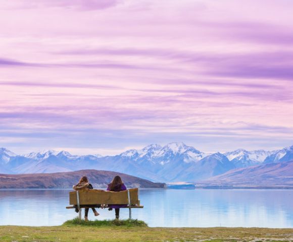 Two people sit on a bench facing a serene lake and snow-capped mountains under a vibrant pink and purple sky, conveying tranquility and awe.