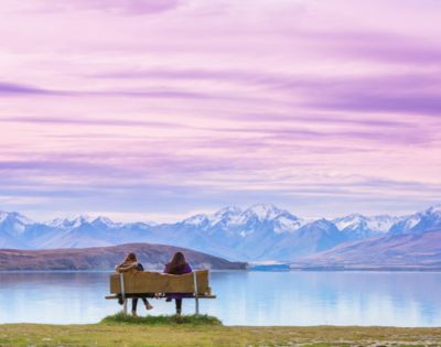 Two people sit on a bench facing a serene lake and snow-capped mountains under a vibrant pink and purple sky, conveying tranquility and awe.