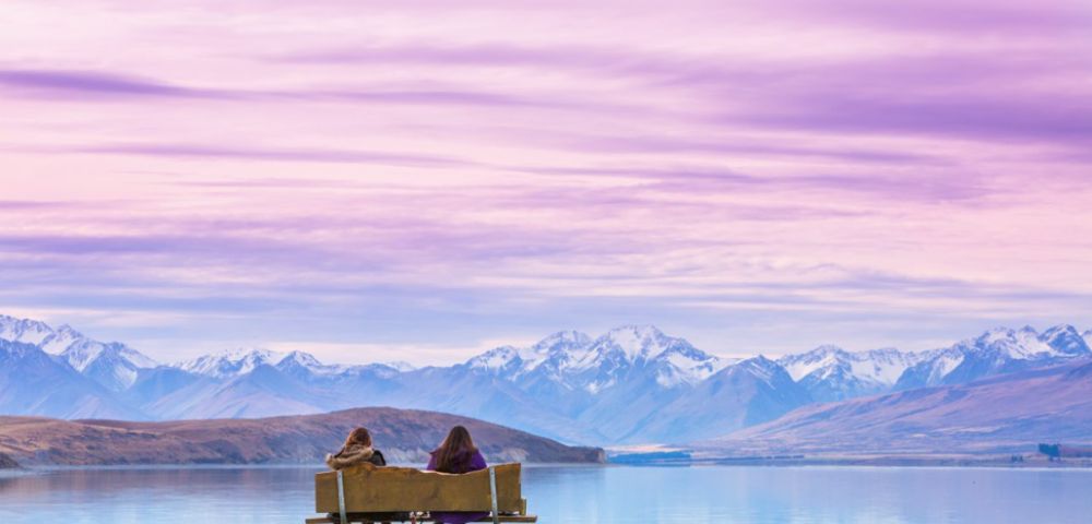 Two people sit on a bench facing a serene lake and snow-capped mountains under a vibrant pink and purple sky, conveying tranquility and awe.