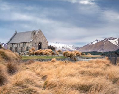 Stone church surrounded by golden grass, set against a backdrop of snow-capped mountains and a cloudy sky, creating a serene and tranquil scene.