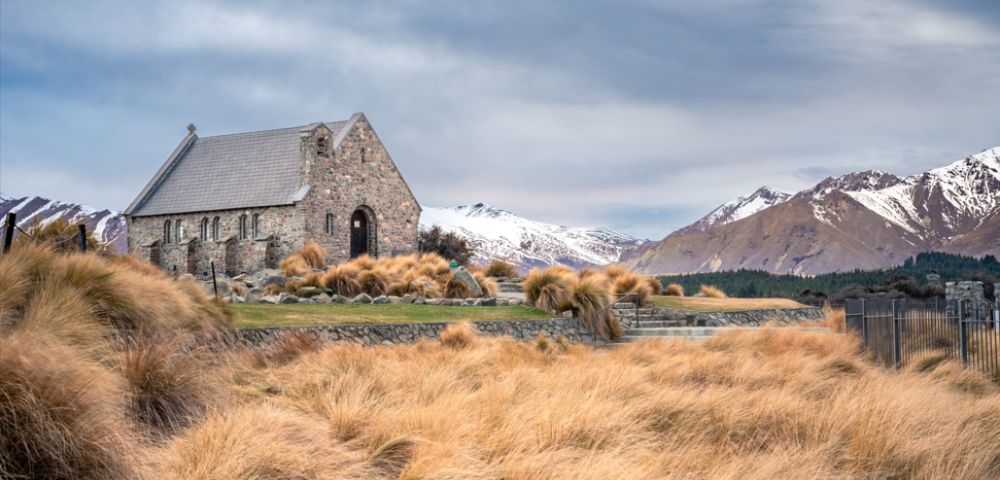Stone church surrounded by golden grass, set against a backdrop of snow-capped mountains and a cloudy sky, creating a serene and tranquil scene.