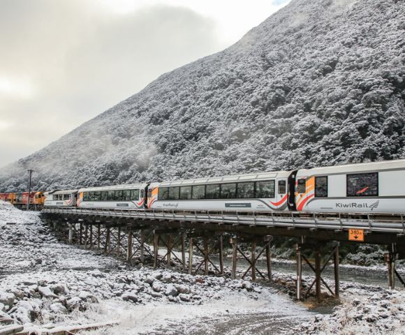 A KiwiRail train crosses a snowy bridge, with a forested mountain in the background. The scene conveys a serene and cold winter atmosphere.