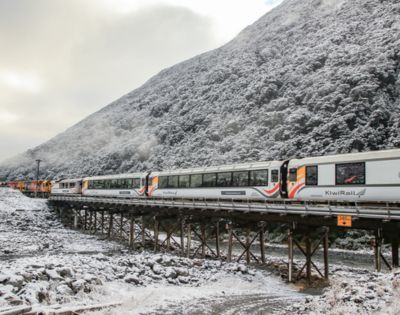 A KiwiRail train crosses a snowy bridge, with a forested mountain in the background. The scene conveys a serene and cold winter atmosphere.
