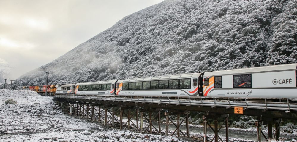 A KiwiRail train crosses a snowy bridge, with a forested mountain in the background. The scene conveys a serene and cold winter atmosphere.